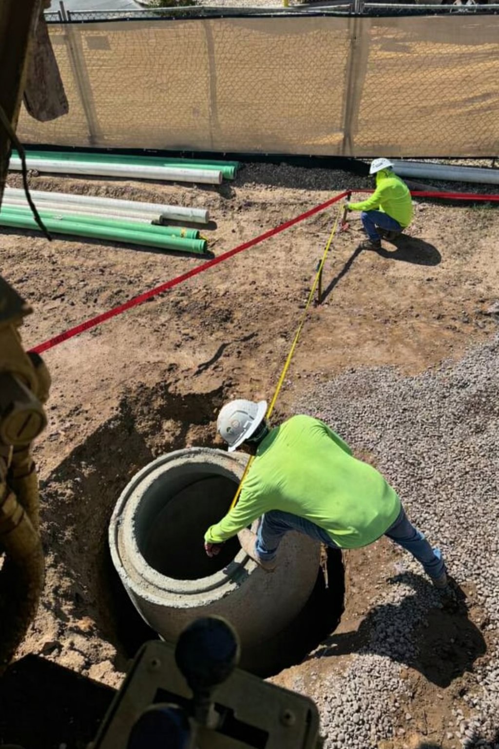 Workers in safety vests and hard hats inspecting a large underground pipe opening at a construction site with materials in background