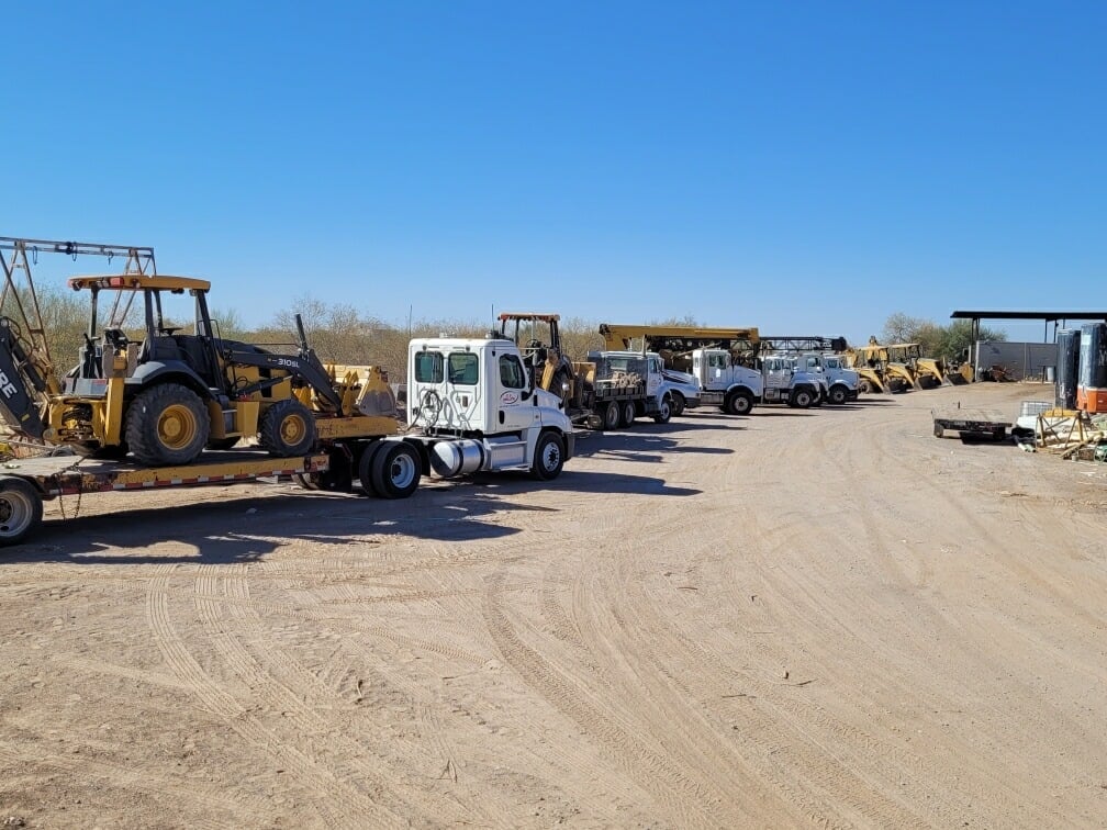 Heavy equipment yard with yellow loader on trailer, white truck, and various machinery parked on sandy lot under clear blue sky